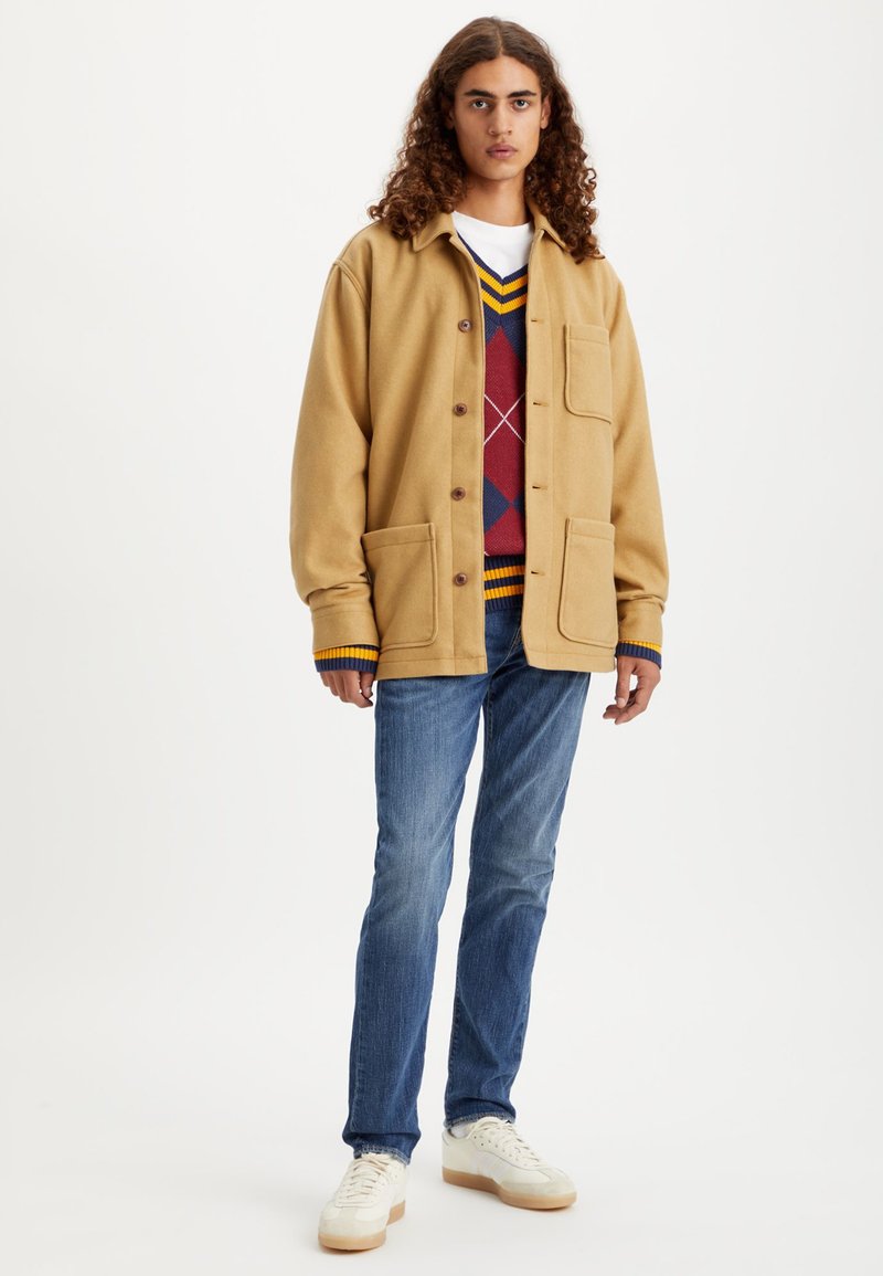 Helle, braune Wollmischjacke mit großen Fronttaschen, die über einem rot-blauen Argyle-Pullover getragen wird. Kombiniert mit blauen Jeans und cremefarbenen Sneakers.
