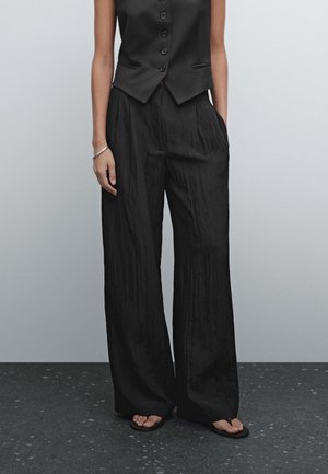 Person iført sort ærmeløs knappet vest, løse sorte brede bukser med tekstur, sorte sandaler og sølvarmbånd, stående på mørkt plettet gulv.