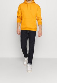 Gul hættetrøje med en frontlomme, kombineret med mørk denimjeans og hvide sneakers. Hættetrøjen har snøre i hætten og ribstrikkede ærmeender.