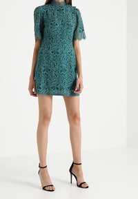 Tyrkysové krajkové šaty s krátkými rukávy, úzkým siluetem, vysokým výstřihem a složitými květinovými vzory, kombinované s černými lodičkami na vysokém podpatku.