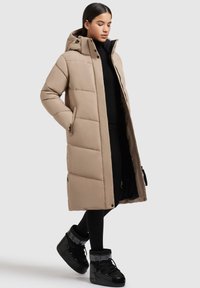 Frau in einem beige langen, gepolsterten Mantel mit Kapuze, schwarzen Leggings und schwarzen Schneestiefeln, die vor einem hellen Hintergrund steht.