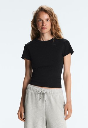 Göndör, szőke hajú nő, aki testhez simuló fekete, rövid ujjú felsőt és világosszürke, megkötős melegítőnadrágot visel, egyszerű háttér előtt.