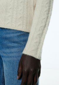 Strickpullover aus wollweißem Material mit strukturiertem Muster und geripptem Saum, kombiniert mit hellblauen Jeans.