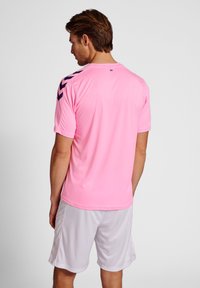 Ljust rosa sporttröja med svarta chevron-accenter på axlarna, i kombination med vita shorts, som har rund hals och korta ärmar.