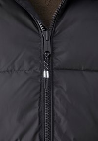 Veste matelassée noire avec fermeture éclair complète à l'avant dotée d'un curseur texturé, finition brillante et coutures horizontales sur tout le corps.