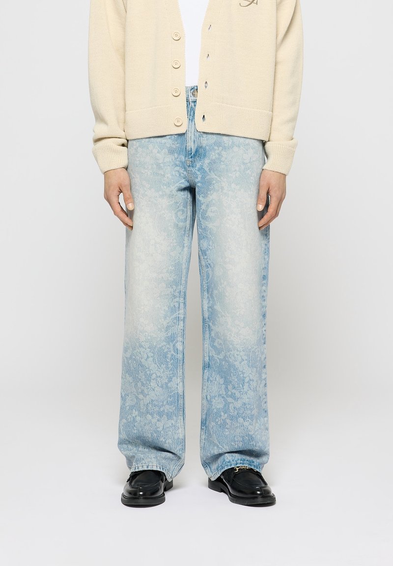 Personne portant un jean large bleu clair à motif floral, un cardigan beige boutonné et des mocassins en cuir noir avec un détail doré.