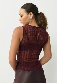 Burgundy, textured sleeveless top with a ruffled hem and keyhole back detail, made of lightweight, sheer fabric.