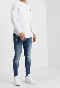 Långärmad vit skjorta med en logotyp på bröstet, tillsammans med slitna blå skinny jeans och vita sneakers. Enkelt, avslappnat design.
