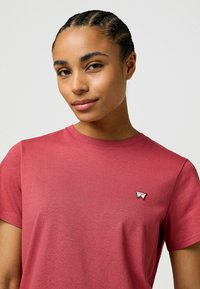 Korte mouwen, rood T-shirt van zachte stof. Heeft een klein wit en rood logo op de linkerkant van de borst. Eenvoudig ontwerp met ronde hals.