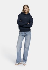 Navy hettegenser med frontlogo, avslappet passform, ribbede mansjetter, kombinert med lyseblå rette jeans og hvite sneakers med grønne striper.