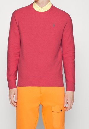 Man wearing a red crew neck sweater over a yellow collared shirt and bright orange pants with side pocket and button flap.