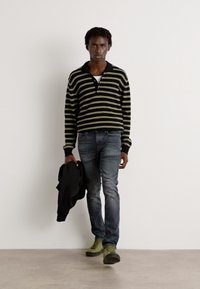 Raido mustikkavihreä raidallinen polo-paita, slim-fit siniset farkut ja oliivinvihreät lenkkarit, vasemmassa kädessä musta takki.