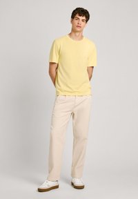Gul bomulds T-shirt med korte ærmer, parret med beige afslappede bukser og hvide sneakers med brune såler. Enkel design, ingen mønstre.