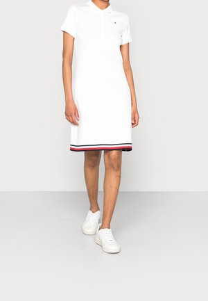 Witte polojurk met een kraag, knoopsluiting en een gestreepte rand in rood en marineblauw aan de onderkant. G gedragen met witte sneakers.