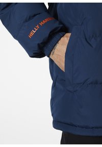 Chaqueta de plumas azul marino con puños elásticos, que presenta un logo naranja de "Helly Hansen" en la manga y un detalle de bolsillo lateral. Textura suave.