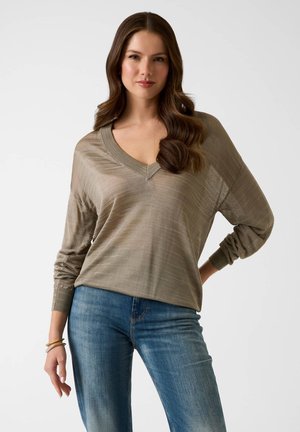 Femme aux longs cheveux ondulés portant un pull ample à col en V taupe et un jean bleu, posant avec une main sur la hanche sur fond blanc.