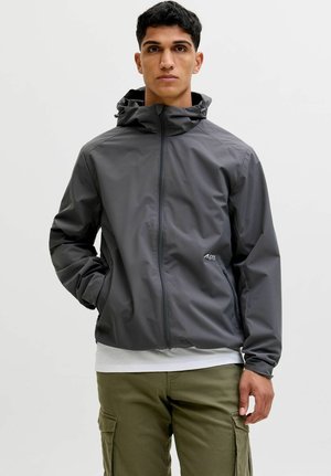 Jonge man met een grijze capuchonvest met ritssluiting en olijfgroene cargobroek, staand met één hand in de zak tegen een effen achtergrond.