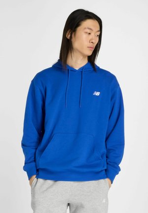 Blauwe hoodie van zacht materiaal, met een voorzak, een capuchon met trekkoord en een klein wit logo op de borst. Grijze broek zichtbaar hieronder.