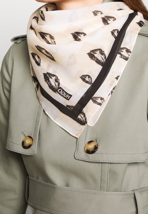 Écharpe beige avec des imprimés de lèvres noires et le texte "HUGO", portée sur un trench-coat vert clair avec de gros boutons en écaille de tortue.