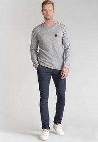 Maglia grigia a maniche lunghe con scollo rotondo e dettaglio a toppa, abbinata a pantaloni slim fit blu navy e sneakers bianche, in piedi davanti a uno sfondo neutro.