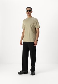 Beige katoenen T-shirt met korte mouwen en een borstzak, gecombineerd met zwarte broek en donker gemusterde sneakers. Accessoires zijn zwarte zonnebril.