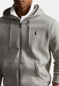 Felpa grigia con zip realizzata in materiale morbido, dotata di una tasca a marsupio, cappuccio con coulisse e un piccolo logo nero sul petto.