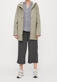 Chaqueta impermeable verde oliva con capucha y cremallera en la parte delantera. Camisa de botones a rayas debajo, combinada con pantalones de pierna ancha en color carbón y zapatillas de deporte.