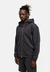 Dunkelgrauer Zip-Hoodie aus strukturiertem Stoff, mit einer Vorderseite-Tasche und verstellbarem Kapuzenzug, kombiniert mit passenden Shorts.