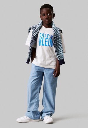 Camiseta blanca con texto azul "CALVIN KLEIN", jeans de color azul claro y zapatillas blancas. Un chaleco a cuadros está drapeado sobre los hombros.