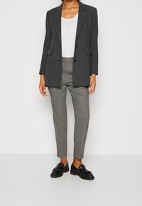Blazer gris croisé porté sur un haut blanc, associé à un pantalon fuselé à carreaux et des mocassins noirs. Tissu lisse avec des motifs subtils.
