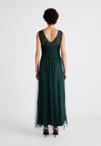 Smaragdgrön klänning med spetsliv och V-ryggdesign. Flödande kjol av genomskinligt material. Klackar synliga längst ner.