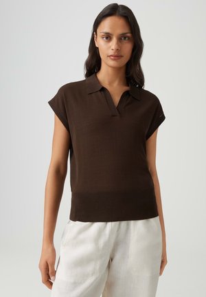 Femme aux longs cheveux foncés portant un polo en maille marron à manches courtes et un pantalon blanc ample sur un fond clair uni.