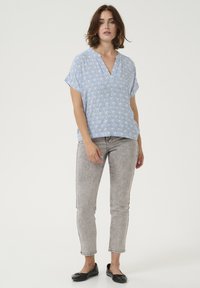 Blouse à manches courtes bleu clair avec un motif géométrique, décolleté en V, associée à un jean gris ajusté et des ballerines noires.