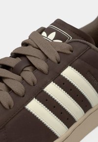 Zapatilla de cuero marrón con rayas beige, cordones marrones texturizados y un logo clásico en la lengüeta. Presenta detalles perforados en los laterales.