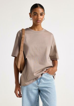 Kvinde iført beige oversize t-shirt, lyseblå jeans og tan skuldertaske, stående med den ene hånd i lommen mod en ensfarvet baggrund.