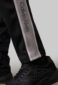 Pantaloni da fitness neri con strisce laterali grigie che presentano il marchio "Calvin Klein", abbinati a sneaker nere testurizzate. Finitura in tessuto liscio.