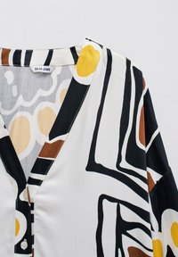 Blouse à motifs avec un décolleté en V, fabriquée en tissu léger. Présente des formes abstraites en noir, blanc, jaune et marron ; comprend un détail de bouton.