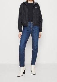 Chaqueta negra con cremallera y capucha gris, combinada con jeans de mezclilla azules y botas blancas hasta el tobillo. La chaqueta presenta un logo en el pecho.