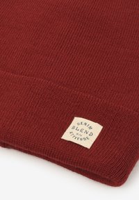 Berretto lavorato a maglia rossa con texture a coste e un'etichetta quadrata con la scritta "DENIM BLEND with ATTITUDE" in lettere nere.