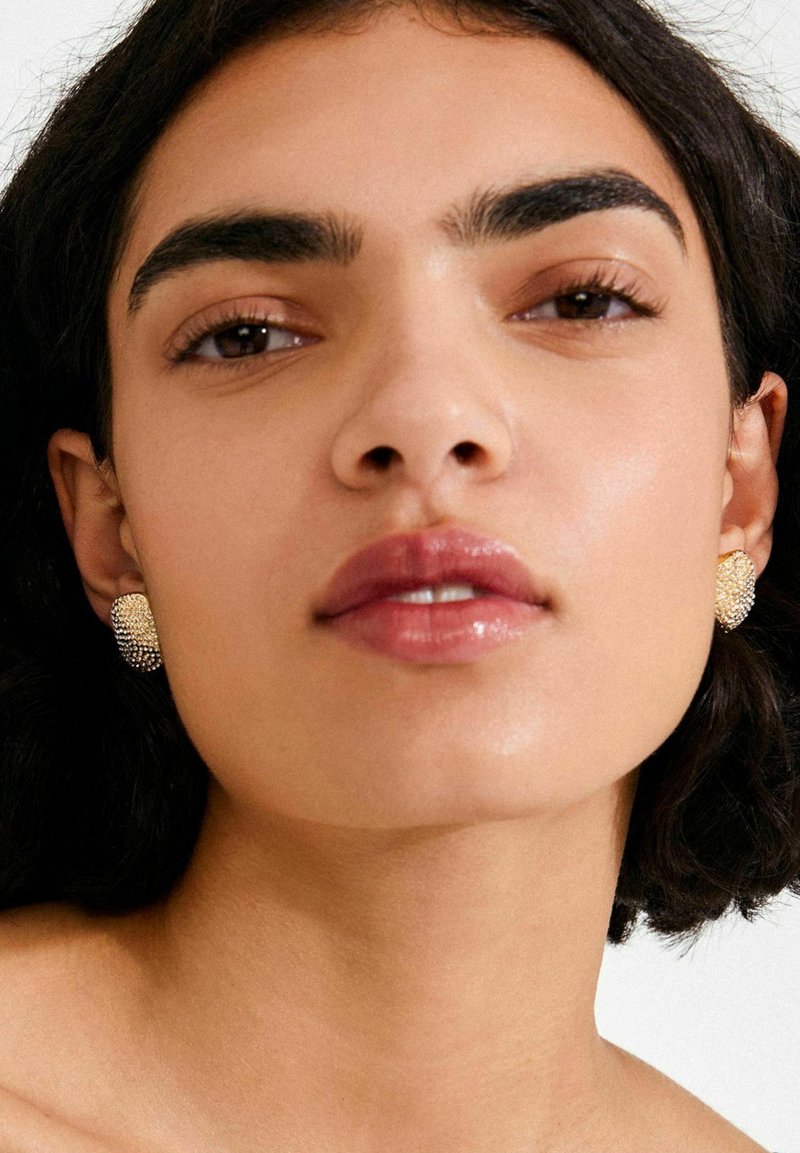 Zelta tonī piespraužamas auskari ar teksturētu virsmu un apļa dizainu, attēloti modeļa ar tumšiem matiem un dabīgu grimu.