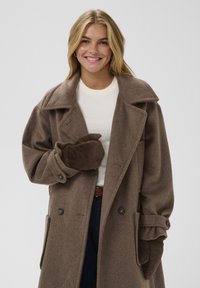 Brūns, divu rindu vilnas mēteli ar lielu apkakli, platām piedurknēm un sānu kabatām. Zem tā valkāts gaišs d�žemperis, roka dzeltenā krāsā atbilstošā brūnā cimdā.