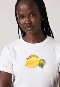 Tricou din bumbac alb, cu un grafism de lămâie galbenă, frunze verzi și o etichetă roșie pe față. Mâneci scurte, decolteu rotund.