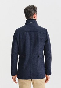 Granatowy płaszcz z wełny, z wysokim kołnierzem, teksturowanym materiałem i detalami z tyłu, w tym dwoma guzikami oraz eleganckim fasonem.