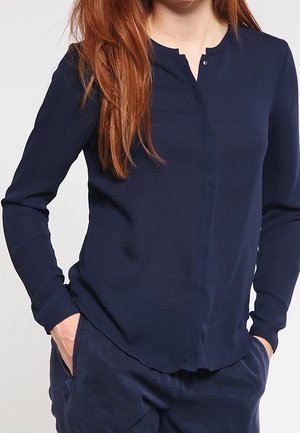 Dunkelblaue Langarmbluse mit Mandarin-Kragen, Knopfleiste und weichem Stoff. Kombiniert mit dunklen Hosen, die eine lässige Passform zeigen.