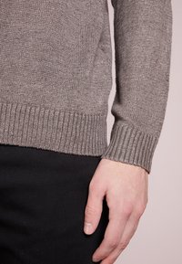 Main reposant le long du corps, portant un pull en maille marron avec des poignets côtelés et un pantalon noir, sur un fond neutre.