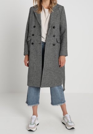 Sort og hvid houndstooth lang frakke med dobbeltbrystede knapper og nedskårne kraver, parret med cropped blå jeans og sporty sneakers.