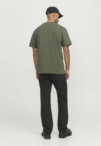 Camiseta de color verde oliva, de manga corta y ajuste relajado, combinada con pantalones negros de pierna recta y zapatos negros, vista desde la parte de atrás.