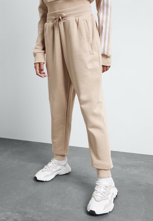 Personne portant un pantalon de survêtement beige Adidas avec taille à cordon et des baskets blanches, debout sur un sol gris contre un mur blanc.
