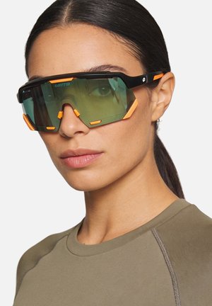 Donna con capelli scuri raccolti, indossa occhiali sportivi oversize neri e arancioni e una maglietta verde oliva, rivolta leggermente verso sinistra.