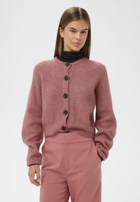 Egy nő rózsaszín gombos kötött kardigánt visel, amely alatt fekete garbót hord, magas derekú rózsaszín nadrággal párosítva, kezével a zsebében.